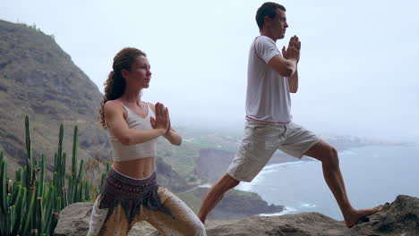 Positioned-on-a-cliff's-brink,-a-man-and-woman-inhale-the-sea-air,-their-hands-raised-during-a-yoga-session-with-the-ocean-view-before-them